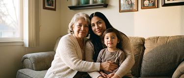 Three generations of women—grandmother, mother, and young daughter—sitting close on a beige sofa in a living room.