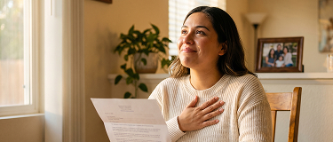 Woman seated at a table near a window, holding a letter to her chest and looking upward with a hopeful expression.